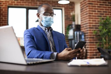 İş kıyafetleri içinde siyah bir adam, yüz maskesi takıyor ve sosyal medyayı cep telefonundan kontrol ediyor. Erkek girişimci dizüstü bilgisayarla masada oturuyor, cep telefonu cihazını tutuyor ve çalışma programını ofisinde planlıyor..