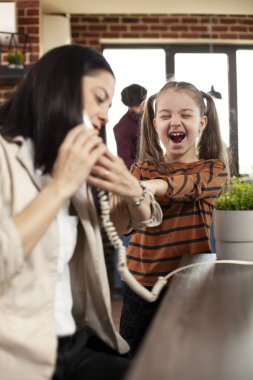 Modern odada, genç kız koluna sarılıp, çalışma alanındaki dikkatini dağıtırken, bayan çalışanlar telefonda konuşuyorlar. Ofiste çalışırken annesine bağıran küçük beyaz bir kız..