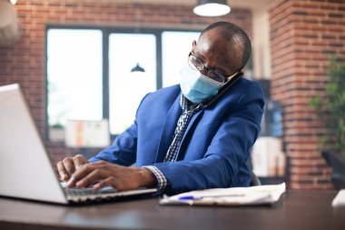 Yüz maskeli bir iş adamı, müşteriyi dinliyor ve laptopta bilgi yazıyor. Erkek yönetici masasında oturmuş, cep telefonuyla konuşuyor ve tuğla duvar ofisindeki kişisel bilgisayarında çalışıyor..