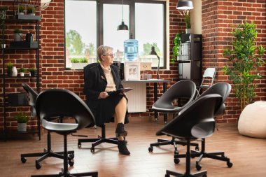 Gözlüklü kıdemli terapist, sandalyelerle çevrili tuğla duvar odasında, elinde pano tutarak rahatça oturuyor. Emekli kadın, parlak ofiste oturmuş hastalarını bekliyor, grup terapisi için hazır..