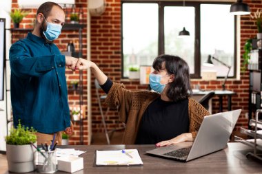 Female analyst and male coworker greet each other with fist bump at desk in startup office. Caucasian colleagues wearing face mask and communicating in safe and cooperative brick wall workspace. clipart