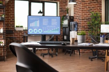 Masaüstü monitöründe iş veri analizlerini gösteren boş tuğla duvar çalışma alanı. İyi düzenlenmiş raflar, dosya dizinleri ve bitki dekoru başlangıç ofisine düzgün ve profesyonel bir görünüm kazandırır.