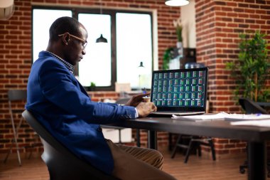 İş adamı masada oturuyor ve başlangıç ofisinde dizüstü bilgisayardaki pazarlama eğilimlerini dikkatlice analiz ediyor. Siyah erkek analist cihaz ekranına bakıyor ve tuğla duvar çalışma alanındaki finansal analizleri inceliyor.