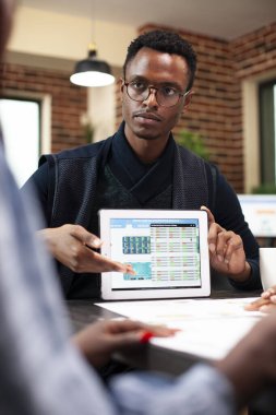 Elinde tablet olan Afrikalı Amerikalı bir adam tuğla duvar ofisindeki bir toplantıda iş piyasası analizini sunuyor. Siyahi erkek yönetici, meslektaşlarına proje güncellemeleri sağlamak için dijital aygıt kullanıyor.
