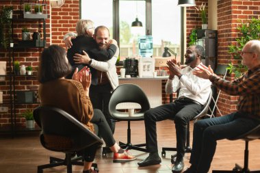 Senior female therapist hugging young man for good mental health progress, claps and smiles at therapy session. Retired counselor embracing male patient for successful recovery, at aa group meeting. clipart