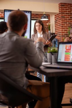 Elinde pano olan beyaz bir kadın, iş planını iş arkadaşlarıyla birlikte yeni açılan ofiste sunuyor. Beyaz kadın yönetici odanın ortasında duruyor ve takımla pazarlama stratejisi hakkında konuşuyor..