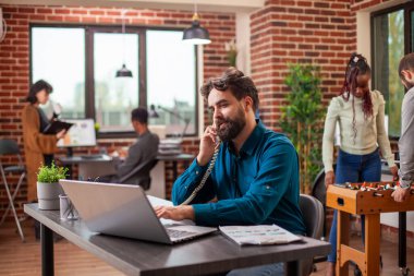 Sakallı yönetici sabit hattan iş arkadaşıyla konuşuyor ve modern ofisteki kişisel bilgisayarda güncellemeleri yazıyor. Erkek girişimci, bir telefon görüşmesi ile dizüstü bilgisayardaki iş detaylarını gözden geçirme arasında birden fazla iş yapıyor.