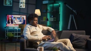 Genç bir yetişkin, dairesinden dizüstü bilgisayarla çalışıyor. Siyah serbest çalışan, rahat bir kanepeden internette gezinirken verimlilik ve rahatlığı harmanlayan görevlere odaklanıyor. Kamera B.