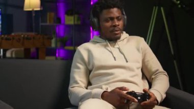 Afro-amerikalı oyuncu evde oturmuş koltuğa oturuyor, oyun oynamak ve boş zamanların tadını çıkarmak için heyecanlanıyor. Dekore edilmiş bir oturma odasında kanepede oturmuş, konsolda video oyunları oynuyor. Kamera B.
