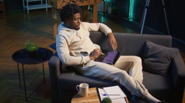 Gülümseyen erkek serbest çalışan modern bir dairede dizüstü bilgisayarla internette geziniyor. Bağımsızlığın yaşam tarzını gösteriyor. Sanal bir çalışma alanında verimlilik, birden fazla iş. Kamera B.