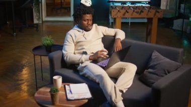 Yetişkin bir adam şık aydınlatılmış dairesinde bir bilgisayarda yazı yazıyor, çevrimiçi işlere karışıyor, birden fazla iş yapıyor ve rahat bir ev ortamında uzaktan çalışmanın esnekliğini kucaklıyor. Kamera B.