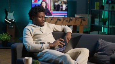 Yetişkin bir adam şık aydınlatılmış dairesinde bir bilgisayarda yazı yazıyor, çevrimiçi işlere karışıyor, birden fazla iş yapıyor ve rahat bir ev ortamında uzaktan çalışmanın esnekliğini kucaklıyor. Kamera A.