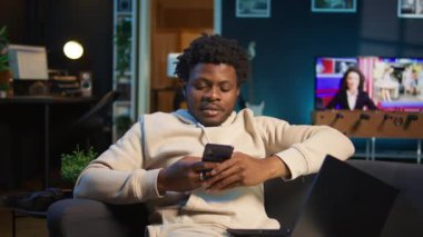 Rahat bir kanepede akıllı telefonuna bakarken gülümseyen rahat bir Afrikalı Amerikalı serbest yazar. Modern ev dekorasyonu şık bir dairede boş zamanların ve çevrimiçi bağlantının altını çizer. Kamera B.