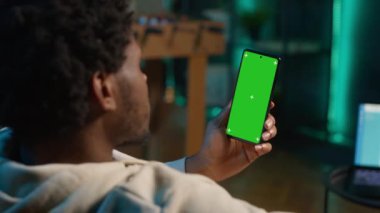 Afrikalı Amerikalı uzaktan kumandalı işçi, modern şık oturma odasındaki kanepede oturarak evinin rahatlığıyla çalışmak için cihazın üzerinde kroma anahtar ekranını kullanıyor. İnternetteki mutlu genç adam. Kamera A.