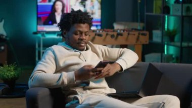 Rahat bir kanepede akıllı telefonuna bakarken gülümseyen rahat bir Afrikalı Amerikalı serbest yazar. Modern ev dekorasyonu şık bir dairede boş zamanların ve çevrimiçi bağlantının altını çizer. Kamera A.