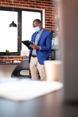 Afrikalı Amerikalı analist tuğla duvar çalışma alanındaki pazar eğilimlerini değerlendirmek için tablet kullanıyor. Yüz maskeli siyahi işadamı, girişimde verimli proje planlaması için dijital aygıt taşıyor.