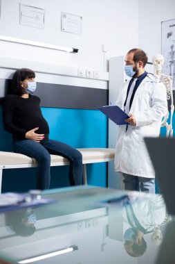 Erkek doğum uzmanı, hamile bir kadının sağlık sorunlarını dinliyor, tedavi güncellemeleri yazıyor ve annelik tavsiyesi veriyor. Doktor hamile bir hastayla muayenehanede doğum öncesi bakımı tartışıyor..