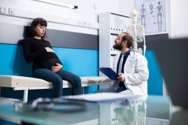 Elinde dosya tutan bir erkek doktor ve hastane yatağında oturan hamile bir kadına danışmanlık yapıyor. Doktor, anne adayının hamilelik muayenesi sırasında karnını okşamasına tıbbi sonuçları iletiyor..