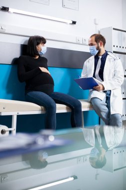 Doğum bakımındaki kadın hasta laboratuvar önlüklü doktorun yanına oturur ve muayene sonuçlarını ve doğum öncesi bakım seçeneklerini sunar. Doktor elinde bir dosya tutuyor ve hamile bayanla birlikte tıbbi talimatları inceliyor..