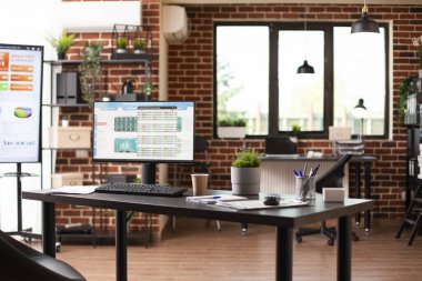 Masaüstü bilgisayarının finansal verilerini iyi organize edilmiş bir masada gösteren kapalı bir başlangıç ofisi. Tuğla duvar çalışma alanı modern teknoloji, raflar ve çalışanların kullanması için hazırlanmış mobilyalar içeriyor..