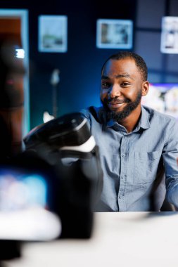 Gülümseyen Afro-Amerikan adam seyircilere VR gözlüklerini sunuyor, profesyonel kamerayla video çekiyor. İçerik yaratıcısı, hayranlarına önermeden önce sanal gerçeklik ürünü deniyor.