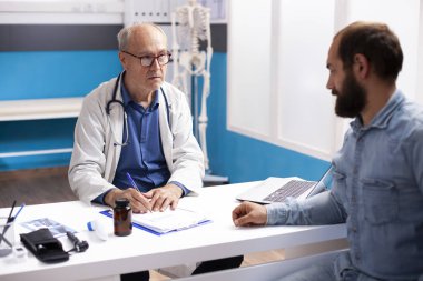 Yaşlı hekim, hastane kontrolü sırasında sakallı hasta bir adamın semptomlarını not alıyor. Emekli doktor, genç erkek hastayla klinik danışmanlığında detayları belgelemek için pano kullanıyor..