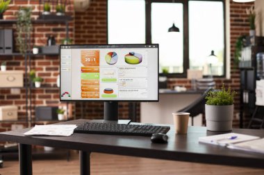 Masaüstü monitörü, not defterleri ve finansal evrakları olan boş tuğla duvar çalışma alanı. Bilgisayar ekranında gösterilen iş analizi verileri ve masadaki belgeler sunum için hazırdır.