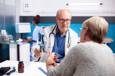 Emekli Kafkasyalı kadın, yaşlı erkek doktorun tıbbi randevu sırasında akciğer radyografisi sonuçlarını tartışmasını dinliyor. Laboratuvar önlüklü yaşlı doktor yaşlı hastaya röntgen sonuçlarını açıklıyor..
