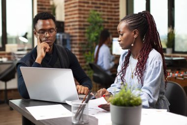 Afrikalı Amerikalı iş arkadaşları dizüstü bilgisayar kullanıyor ve online bilgileri ofis masasındaki evraklarla karşılaştırıyorlar. Siyahi işadamı ve kadın birlikte oturmuş dijital bir cihazla şirket araştırması yapıyorlardı..