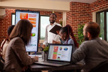 Siyah işadamı monitörün yanında duruyor iş analitik verilerini gösteriyor ve iş arkadaşlarıyla ofis tartışması yapıyor. Tuğla duvar ofisinde verimli toplantı için dijital teknoloji kullanılıyor.