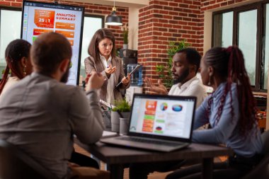 Bir masa etrafında oturan iş arkadaşlarıyla proje fikirlerini sunan bir pano yöneticisi. Meslektaşlar, ortak bir toplantıda strateji oluşturmak için finansal tabloları ve teknolojiyi kullanıyorlar..