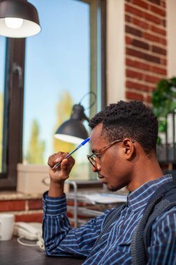 Siyahi girişimci başlangıç ofisinde masada oturuyor, elinde kalem tutuyor ve proje geliştirme için masa başında belgeler okuyor. Profesyonel danışman. Gözlüklü. Duvarda alnına vuruyor..