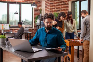 Erkek ofis çalışanı, masadaki dosya ve dosyaları incelemeye odaklanmış. Dizüstü bilgisayar, tuğla duvar çalışma alanında onun yanına yerleştirilmiş. Genç adam masada oturup şirket yönergelerini okuyor..