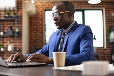 Yönetici masadaki kahveyle dizüstü bilgisayarda çalışıyor, e-postaları ve başlangıç ofisindeki belgeleri inceliyor. İş adamı kişisel bilgisayarında yazı yazıyor, tuğla duvar odasında pazarlama stratejisini araştırıyor..