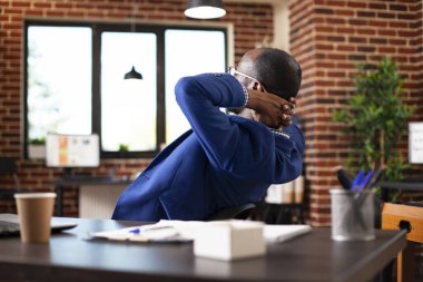 Yönetici masasında dinleniyor, sandalyeye yaslanıyor ve başlangıç ofisinde dinleniyor. Elleri başının arkasında olan siyahi işadamı modern tuğla duvar çalışma alanında dinlenmek için duruyor..