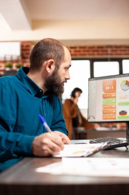 Şirketin araştırma notlarını elinde kalemle ofis masasında okumaya odaklanmış bir işadamı. Sakallı bir erkek analist, masaüstü bilgisayarındaki mali tabloları inceliyor ve yakınlarda bir evrak işi var.