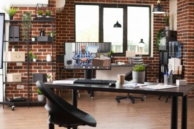 Tekerlekli sandalyedeki iş adamının video çağrısını gösteren bilgisayar ekranlı boş tuğladan bir işyeri. Masaüstü bilgisayarı beyaz adamla boş başlangıç ofisinde devam eden sanal toplantıyı gösteriyor.