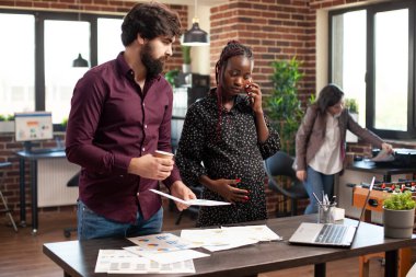 Hamile kadın telefonda konuşuyor ve tuğladan ofis çalışanı bir erkekle dizüstü bilgisayardaki mali tabloları inceliyor. Mobil cihazlı beklenen kadın iş adamıyla proje sunumu hazırlıyor.