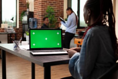 Siyah kadın yakınlarda otururken boş bir model şablonu olan dijital cihaza odaklan. Ofiste çalışan kadın çalışan, masasında yeşil ekranı gösteren bir dizüstü bilgisayarla..