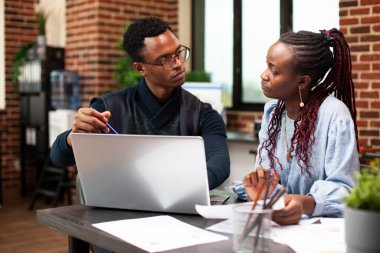 Siyahi erkek yönetici, iş arkadaşıyla yaptığı görüşme sırasında cihazlı ekranı işaret ederek ona güncel iş analizlerini gösteriyor. Bilgisayarlı bir iş adamı, tuğla duvar ofisindeki kadın çalışanıyla konuşuyor..