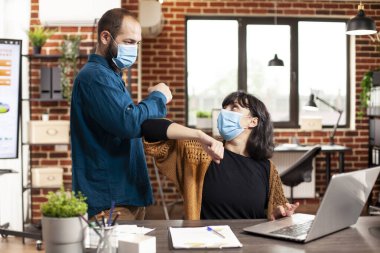 Masada çalışan iş kadını, erkek iş arkadaşı onu tuğladan duvar ofisinde dirsekle selamlıyor. İş adamı ve bayan iş arkadaşı yüz maskesi takıyor ve yeni başlayan şirkette güvenliği sağlıyor..