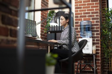 Başlangıç ofisinde masaüstü bilgisayarı üzerinde çalışan profesyonel bir kadın, pazarlama ve eğilimler üzerine gayretli bir araştırma yürütüyor. Masaya oturmuş bilgisayar ekranına odaklanmış genç bir kadın verileri inceliyor.