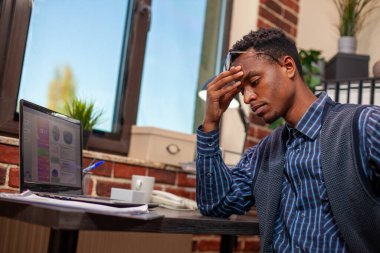 African American entrepreneur sits tiredly at desk, holding his head as he analyzes business project plans. Black male manager looking drained after demanding day at work in startup office. clipart