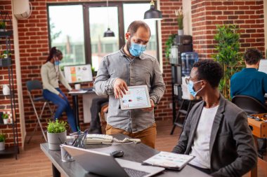 Beyaz bir adam güvenli ofis ortamında siyahi meslektaşıyla konuşurken tablet üzerinde veri sunuyor. Masada çalışan erkek iş arkadaşları. İkisi de modern çalışma alanında maske takıyor..