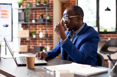 Siyah işadamı dizüstü bilgisayarla masada oturuyor ve proje fikirlerini düşünüyor. Afrika kökenli Amerikalı yönetici, başlangıç ofisinde çalışırken alnına dolma kalemle dokunuyor..