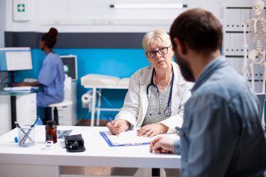 Emekli kadın doktor, arka planda çalışan genç erkek hastaya danışmanlık yapıyor. Yaşlı doktor sakallı adamı dinliyor ve tıbbi belirtilerini yazıyor..