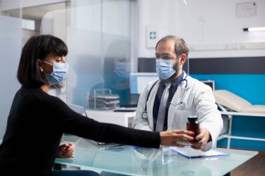 Doktor ağrı kesicileri ve sağlık desteğini açıklarken genç bir kadın klinik randevusunda oturuyor. Doktor, masadaki koruyucu ekranla kadın hastaya reçeteli ilaç veriyor..