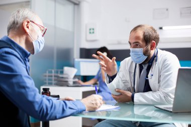 Emekli adam, klinikte yüz maskesi takan genel doktorla tedavi planı hakkında görüşüyor. Eski bir hasta panoya yazı yazıyor, sağlık kontrolü için nazik bir doktordan yardım alıyor..