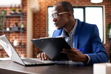 Gözlüklü bir işadamı şirket araştırma belgelerini tutuyor ve dizüstü bilgisayardaki verileri kontrol ediyor. Ofis masasında oturan erkek analist, panodan gelen bilgilerle birlikte cihazdaki iş analizlerini güncelliyor..