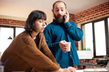 Erkek çalışan müşteriyle telefonda konuşuyor ve iş projesi sunumunu iş arkadaşıyla gözden geçiriyor. Kadın meslektaşı şirket araştırma belgelerinin incelenmesine yardım ederken, iş adamı mobil cihazla konuşuyor.